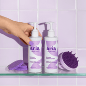 Two bottles of Aria Haircare blonde shampoo and conditioner on a glass shelf with purple sponges and a brush against a tiled wall background.