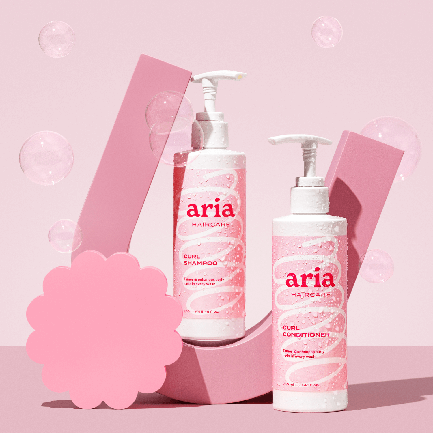 Aria haircare curl shampoo and conditioner bottles on a pink background with bubbles.