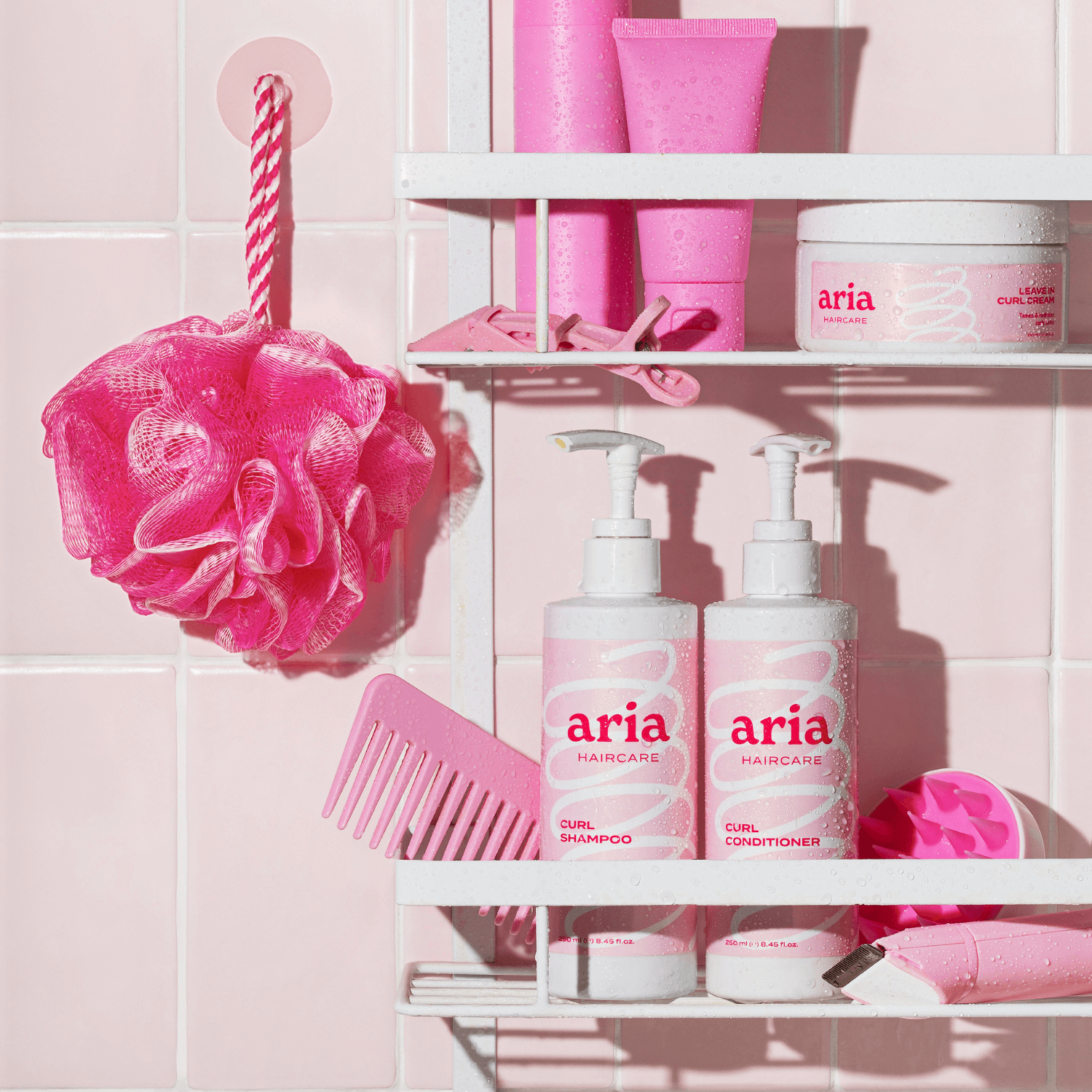 Bathroom shelf with pink aria haircare curl shampoo, conditioner and curl cream products against a pink tiled wall.